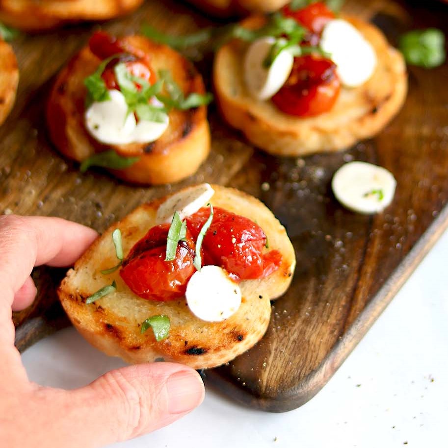 two roasted cherry tomatoes with slices of fresh mozzarella on slice of baquette in focus being held by a hand, others on wooden cutting board slightly blurry in background