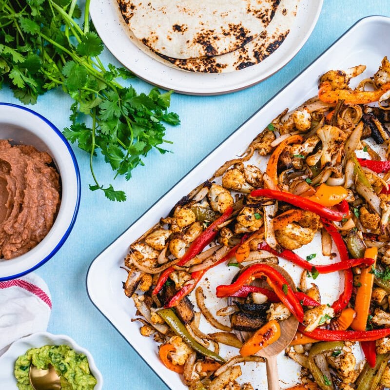 sheet pan with roasted red and yellow capsicum, cauliflower florets and mushrooms, next to a bowl of refried beans, plate of tortillas and bowl of guag, with fresh coriander
