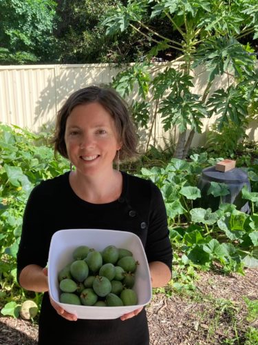 Kylie holding a container of home grown feijoas with her lush home garden and compost in background