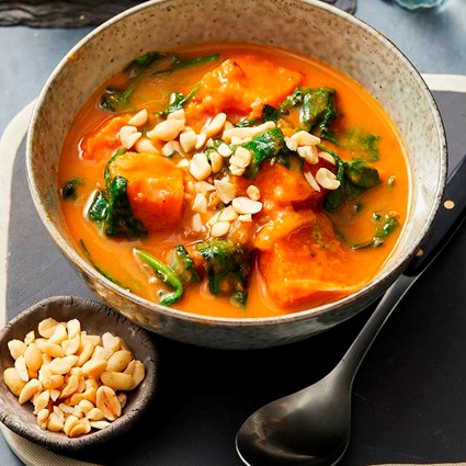 Large bowl with orange curry, large chunks of sweet potato, wilted green leaves and peanuts on top