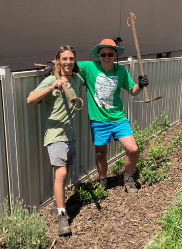 A man in green shirt with sunglasses and hat has his arm around younger man. Both are holding garden picks and smiling. It is sunny and looks warm