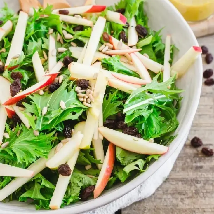 fresh green mizuna leaves with apple matchsticks and sultanas in white bowl