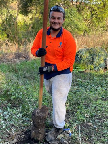 Young man standing in market garden holding bamboo pole vertically. He is smiling at the camera and wearing an orange high vis shirf and light pants.