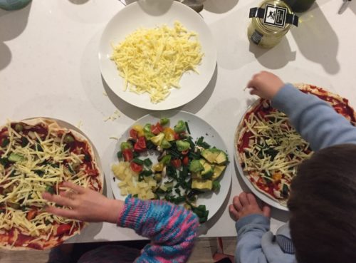 kids making pizzas: top view with plate of cheese and plate of chopped veggies in middle, with 2 kids on either side assembling ingredients on pizza bases